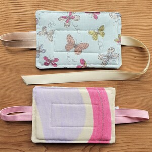 May include: Two fabric hot water bottles with straps. The top one is light blue with a butterfly pattern and a cream-coloured strap. The bottom one is purple and pink with a cream-coloured strap.