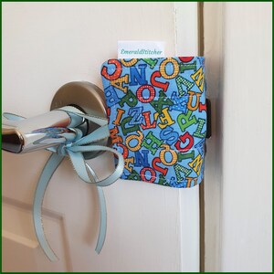 May include: A blue fabric doorstop with colourful letters printed on it. The doorstop is attached to a white door with a silver door handle. A blue ribbon is tied around the door handle.