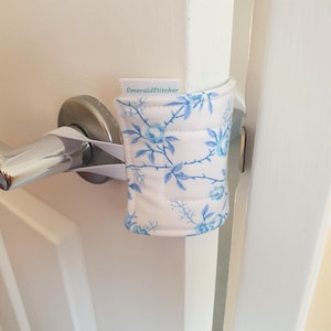 May include: A white doorstop with a blue floral pattern and the text "EmeraldStitcher" on the top. The doorstop is attached to a white door handle with a white strap.