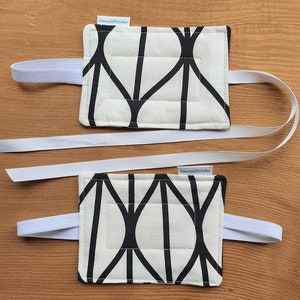 May include: Two black and white patterned oven mitts with white elastic straps. The mitts have the text "Emerald Stitcher" printed on them.