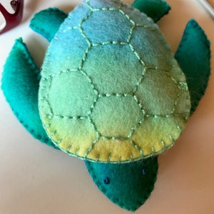 Puede incluir: Una tortuga de mar de fieltro verde y azul con un caparazón cosido. El caparazón es un degradado de colores que va del azul claro al verde al amarillo.