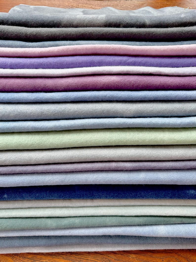 May include: A stack of 18 fabric squares in various shades of blue, green, purple, and gray. The fabric is soft and has a slightly textured surface.