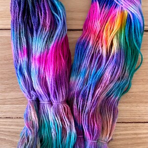 May include: A skein of hand-dyed yarn in shades of purple, blue, green, pink, and yellow. The yarn is arranged in a loose circle, showing the colour variations.