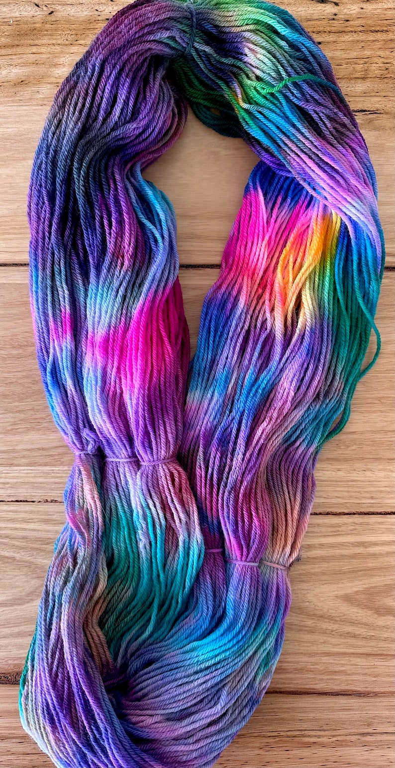 May include: A skein of hand-dyed yarn in shades of purple, blue, green, pink, and yellow. The yarn is arranged in a loose circle, showing the colour variations.