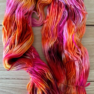 May include: A skein of hand-dyed yarn in shades of pink, orange, and yellow. The yarn is arranged in a loose coil.
