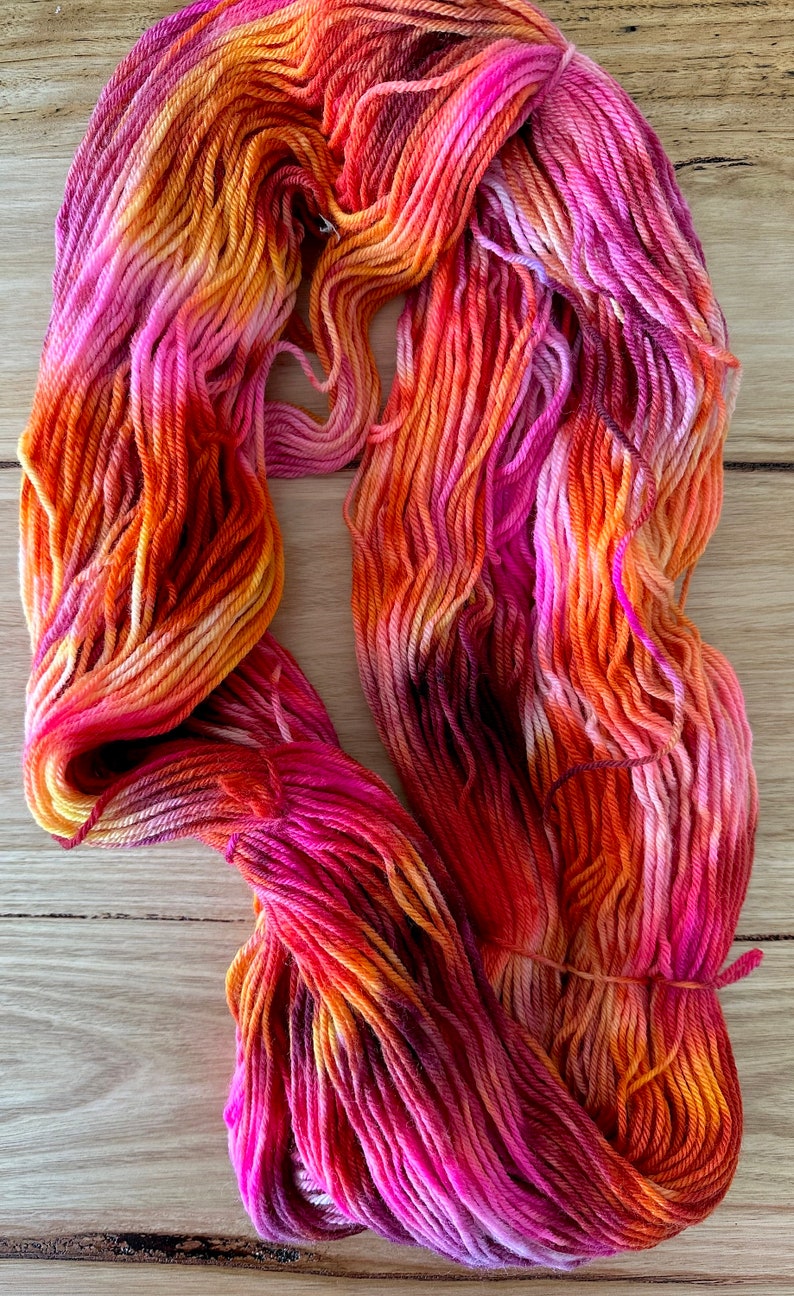 May include: A skein of hand-dyed yarn in shades of pink, orange, and yellow. The yarn is arranged in a loose coil.