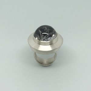 May include: A silver ring with a black and white gemstone set in a dome-shaped bezel. The ring has a wide band and a raised, circular setting.