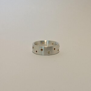 May include: A silver ring with a matte finish. The ring is adorned with small, evenly spaced, amber-colored gemstones. The design features a series of circular inlays, creating a unique and modern aesthetic. This piece of jewelry is suitable for everyday wear.