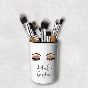 May include: White ceramic makeup brush holder with a gold glitter eye design and the text "Rachel's Brushes".