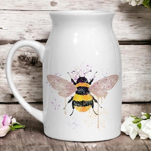 Könnte beinhalten: Weiße Keramiktasse mit einer Aquarell-Illustration einer Hummel mit gelben und schwarzen Streifen und braunen Flügeln. Die Hummel ist von violetten und gelben Aquarellspritzer umgeben.