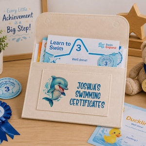 May include: A cream-colored felt document holder featuring a blue dolphin graphic and the text JOSHUA'S SWIMMING CERTIFICATES. The holder contains a Learn to Swim 3 certificate, accompanied by a blue rosette, a circular badge, and a Duckling certificate.