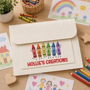 May include: A cream-colored felt portfolio case featuring a graphic of six colorful cartoon crayons with faces and the text "HOLLIE'S CREATIONS" in red lettering. The case has a flap closure and is surrounded by various children's drawings.
