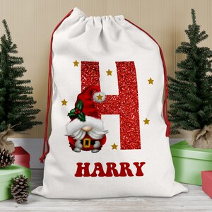 May include: White Christmas gift bag with a red glitter letter H and a cartoon gnome with a red hat. The bag has gold stars and the name Harry printed in red.