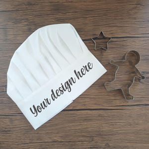 May include: A white chef's hat with the text "Your design here" in black script. Beside the hat are two metal cookie cutters, one shaped like a star and the other like a gingerbread person. The items are on a wooden surface.