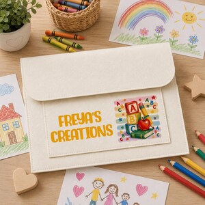 May include: A cream-colored felt document folder featuring the text "FREYA'S CREATIONS" in yellow letters next to a graphic of alphabet blocks, a red apple, and a pencil. The folder is surrounded by colorful children's drawings and crayons.