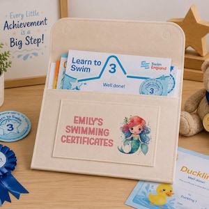 May include: A beige felt document folder with a flap, featuring the text "EMILY'S SWIMMING CERTIFICATES" and a mermaid illustration. Inside are "Learn to Swim 3" certificates. Nearby are a blue rosette, a circular badge, and a teddy bear with a medal.