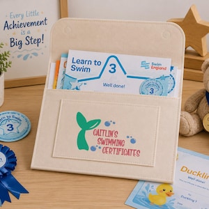 May include: A cream-colored felt document folder with a flap closure, featuring a green mermaid tail graphic and the text "Caitlin's Swimming Certificates" printed on the front pocket, containing several blue and white swimming achievement certificates.