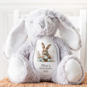 May include: A gray plush bunny rabbit with a white patch on each ear. The bunny is holding a blue egg and sitting in a nest with other blue eggs. The bunny has a white patch on its chest with the text "Oliver's First Easter 2025".