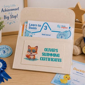 May include: A beige felt document holder featuring a stitched rectangular pocket with a cartoon fox wearing goggles and the text Oliver's Swimming Certificates. The holder contains a blue and white Learn to Swim 3 certificate from Swim England.