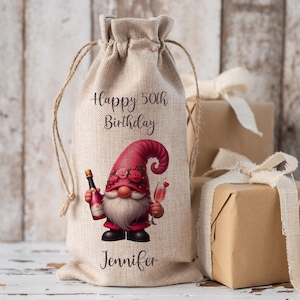 May include: A beige burlap gift bag with a drawstring. The bag has a colourful illustration of a gnome holding a bottle and a glass of champagne. Text on the bag reads "Happy 50th Birthday Jennifer."