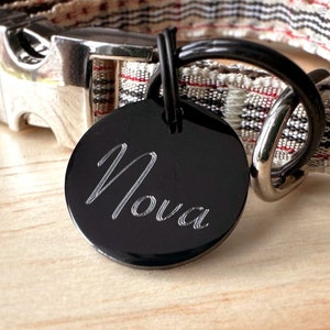 May include: A black engraved dog tag with the name "Nova" on it, attached to a white and black striped dog collar.