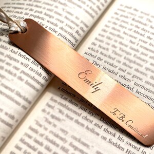 Peut inclure: Un marque-page en métal de couleur cuivrée, gravé du nom "Emily" et de la phrase "To Be Continued". Le marque-page rectangulaire, avec une extrémité arrondie et un trou pour un ruban, est posé sur les pages ouvertes d'un livre.