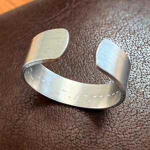 May include: A silver metal bracelet with a smooth, polished finish. The bracelet is open and has a simple design. The inside of the bracelet is engraved with a series of small lines.
