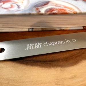 May include: A silver metal bookmark with the text "chapters in ♡" and a tally mark design. The bookmark has a leather cord attached and is resting on a wooden surface next to a book.