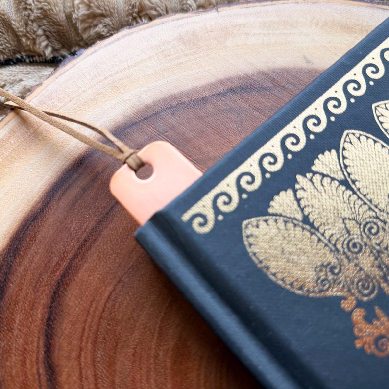 May include: A peach-colored wooden bookmark with a leather cord is inserted into a dark blue book with gold decorative patterns. The book is resting on a wooden surface.