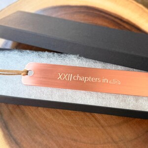 May include: A copper-colored metal bookmark with the text "XXII chapters in" and a small design. The bookmark is in a black gift box with a leather cord attached. The box is on a wooden surface.