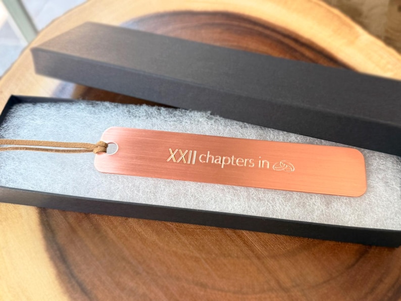 May include: A copper-colored metal bookmark with the text "XXII chapters in" and a small design. The bookmark is in a black gift box with a leather cord attached. The box is on a wooden surface.