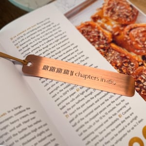 May include: A copper-colored metal bookmark with the text "chapters in" and a leather cord. The bookmark is lying on an open cookbook, with a recipe for cinnamon rolls visible.