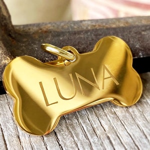 May include: A polished gold-coloured dog tag shaped like a bone, with the name "LUNA" engraved on it. The tag has a small loop for attaching to a collar. The background is a blurred view of a wooden surface.