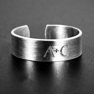 May include: A silver-toned adjustable ring with a brushed metal finish. The ring features the engraved letters "A + C" on the front. The ring is open at the top, allowing for size adjustment. The ring is set against a dark background.