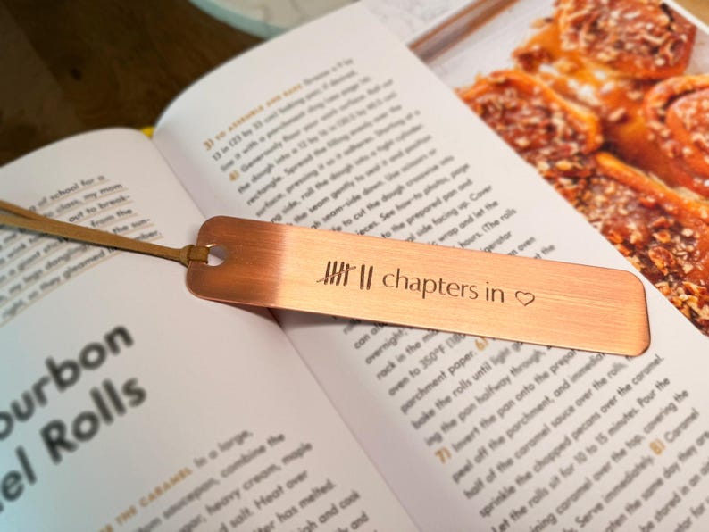 May include: A copper bookmark with the text "chapters in" and a heart symbol, resting on an open cookbook. The bookmark has a leather cord attached. The book is open to a recipe for bourbon rolls.