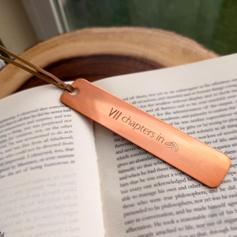 May include: A copper bookmark with a leather cord rests on an open book. The bookmark has the text "VII chapters in" and a small design. The bookmark is rectangular with rounded edges and a hole for the cord.