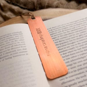 May include: A copper-colored bookmark with the text "XXII chapters in" is placed in an open book. The bookmark has a small hole at the top with a brown string attached. The book's pages are white with black text.