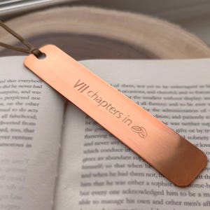 May include: A copper-colored metal bookmark with the text "VII chapters in" engraved on it. The bookmark has a small hole at the top with a brown leather cord attached. The bookmark is resting on an open book.
