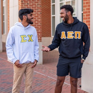 May include: Two people wearing hoodies with Greek letters on them. The person on the left is wearing a light grey hoodie with the letters "ΣΧ" in yellow and blue. The person on the right is wearing a black hoodie with the letters "ΑΕΠ" in yellow and blue.