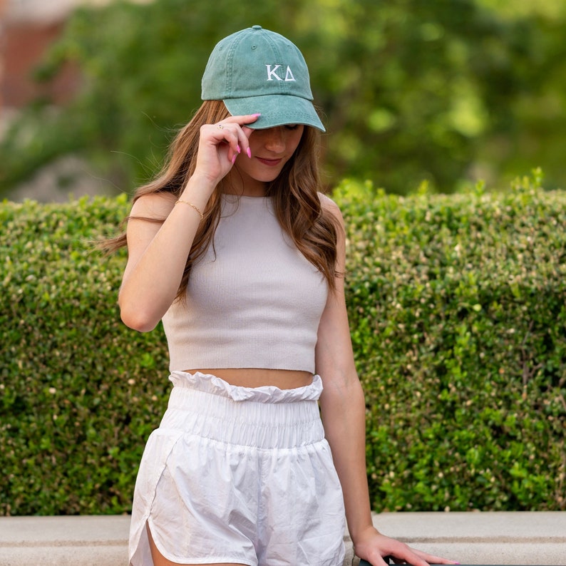 May include: A green baseball cap with white embroidered letters "KA" on the front. The cap is worn by a person with long brown hair. The person is wearing a white tank top and white shorts.