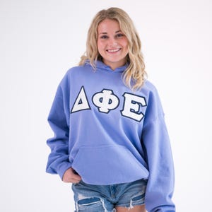 May include: A periwinkle blue hooded sweatshirt with a kangaroo pocket. The sweatshirt features the Greek letters ΔΦΕ in white with black trim across the chest. The model is wearing denim shorts.