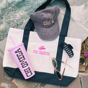 Peut inclure: Un sac fourre-tout blanc avec des sangles noires et un motif brod&eacute; rose qui dit "Chi Omega". Le sac est pos&eacute; sur une surface brune et contient une casquette de baseball grise avec le texte "Chi O" brod&eacute; dessus, un gobelet rose avec le texte "Chi Omega" dessus, une pince &agrave; cheveux noire et une paire de lunettes de soleil brunes.