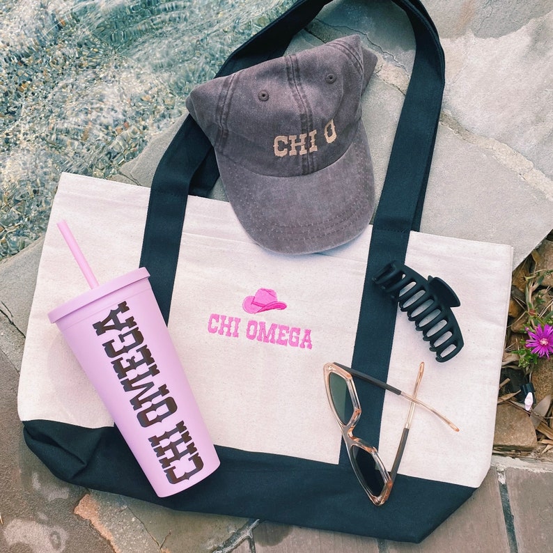 Peut inclure: Un sac fourre-tout blanc avec des sangles noires et un motif brod&eacute; rose qui dit "Chi Omega". Le sac est pos&eacute; sur une surface brune et contient une casquette de baseball grise avec le texte "Chi O" brod&eacute; dessus, un gobelet rose avec le texte "Chi Omega" dessus, une pince &agrave; cheveux noire et une paire de lunettes de soleil brunes.