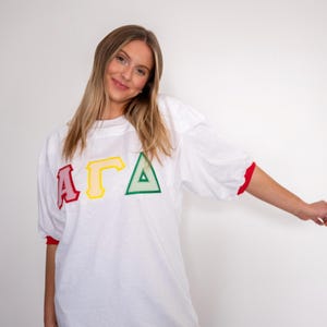 May include: A white short-sleeved jersey featuring the Greek letters Alpha, Gamma, and Delta in red, yellow, and green. The letters are appliqued with a striped pattern. The shirt has a crew neckline and red trim on the sleeve cuffs.