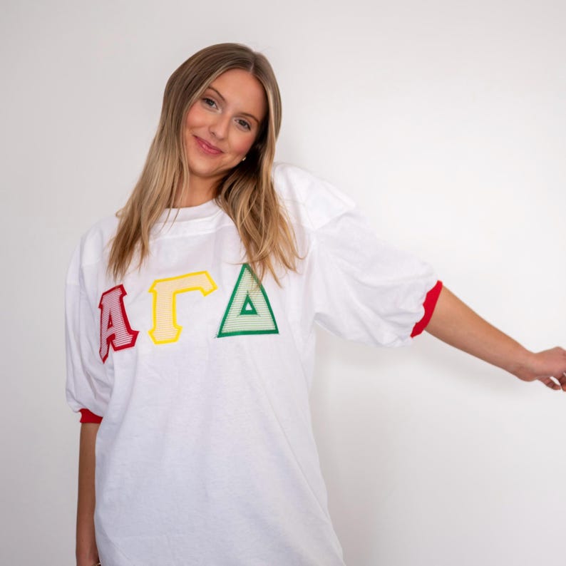 May include: A white short-sleeved jersey featuring the Greek letters Alpha, Gamma, and Delta in red, yellow, and green. The letters are appliqued with a striped pattern. The shirt has a crew neckline and red trim on the sleeve cuffs.