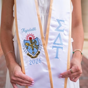May include: White graduation sash with light blue embroidered Greek letters and the name "Kyndall". The sash has a gold trim and a crest with the year "2026".