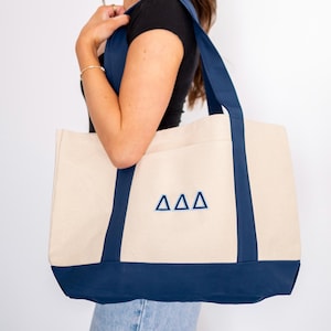 May include: A large, white canvas tote bag with navy blue trim and handles. The bag has a blue embroidered Greek letter design that reads "ΔΔΔ".
