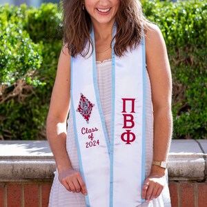 May include: A white graduation stole with blue trim, featuring a red and white crest and the text "Class of 2024" and "ΠΒΦ".