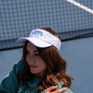 May include: A white visor with a blue embroidered "KAPPA" logo. The visor is worn by a person with long brown hair.