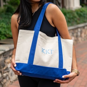 May include: A white canvas tote bag with blue handles and a blue bottom. The bag has the letters "KKT" embroidered on the front in blue thread.